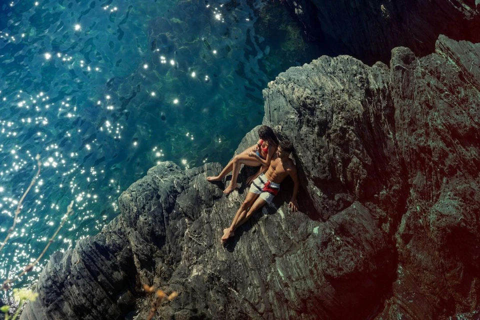 Couple relaxes on rocky outcrop above sparkling turquoise water, enjoying the sunshine and scenic view. Summer vacation travel concept.