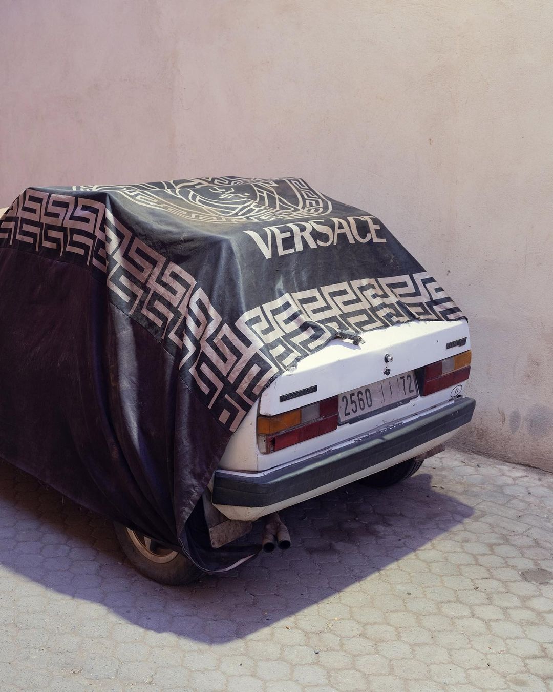 A car parked outside, partially covered with a black Versace-branded tarp, casting a shadow on the patterned concrete.