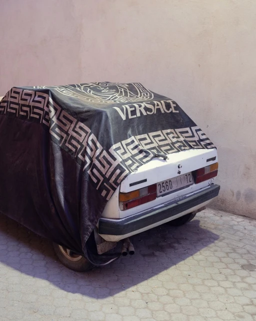 A car parked outside, partially covered with a black Versace-branded tarp, casting a shadow on the patterned concrete.
