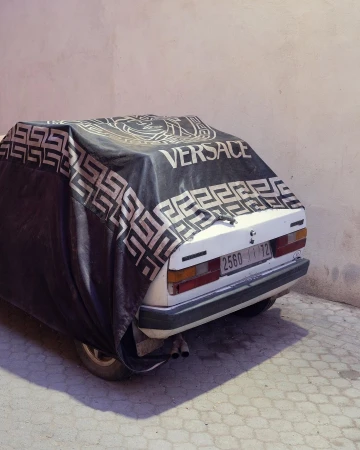 A car parked outside, partially covered with a black Versace-branded tarp, casting a shadow on the patterned concrete.