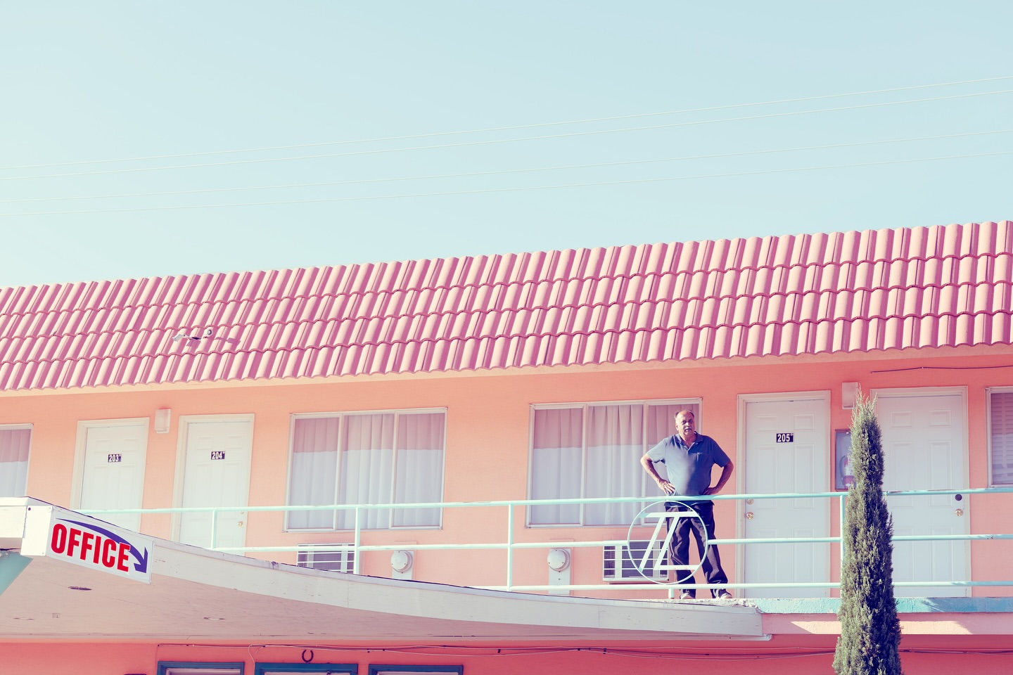 Man stands on motel balcony, looking at the camera. The building is peach-colored with a red roof and an 'Office' sign.