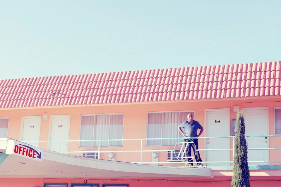 Man stands on motel balcony, looking at the camera. The building is peach-colored with a red roof and an 'Office' sign.