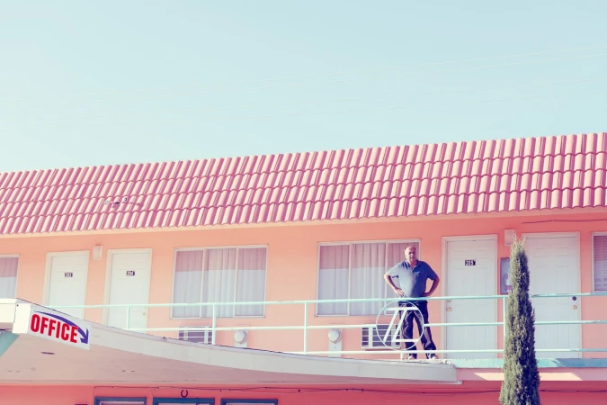 Man stands on motel balcony, looking at the camera. The building is peach-colored with a red roof and an 'Office' sign.