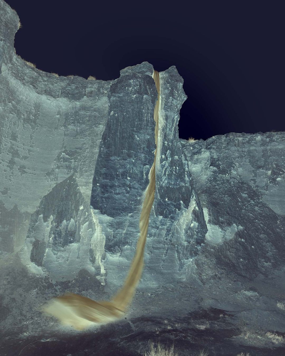 Landscape photograph of a tall rock formation with a flowing waterfall under a dark sky, showcasing natural beauty and geological formations.