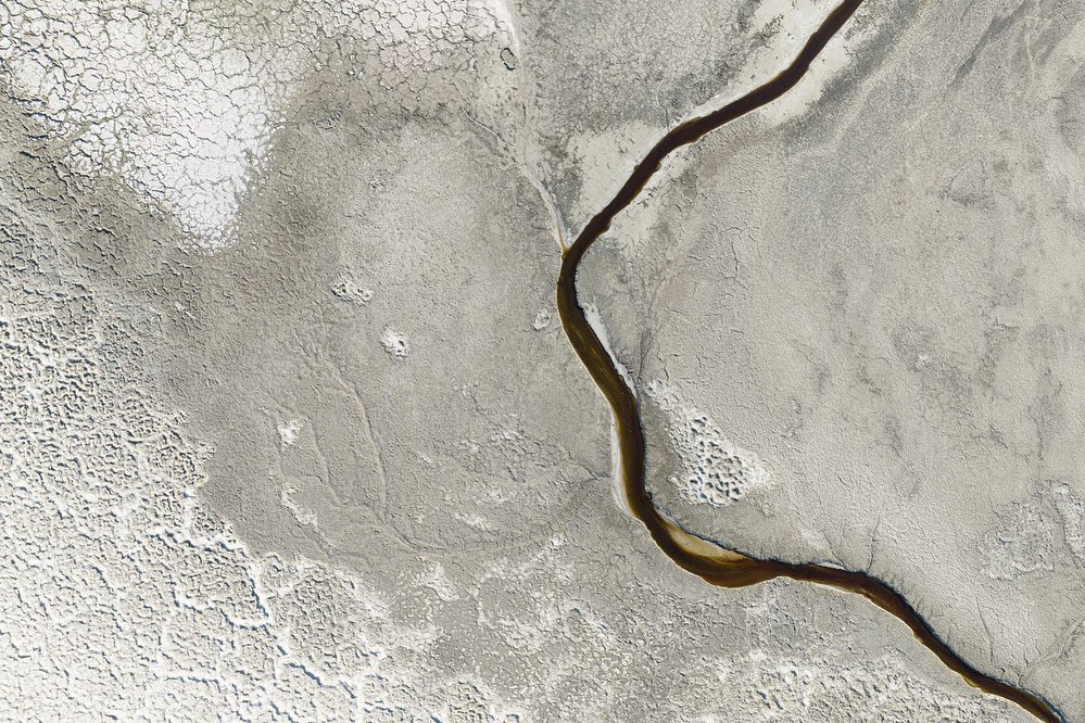Aerial view of cracked earth terrain with a winding stream, showcasing patterns of drought and erosion in a neutral color palette.