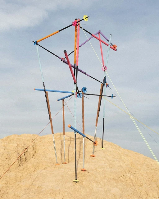 Abstract art installation on a sand dune against a blue sky. The sculpture features colorful poles and geometric shapes. 