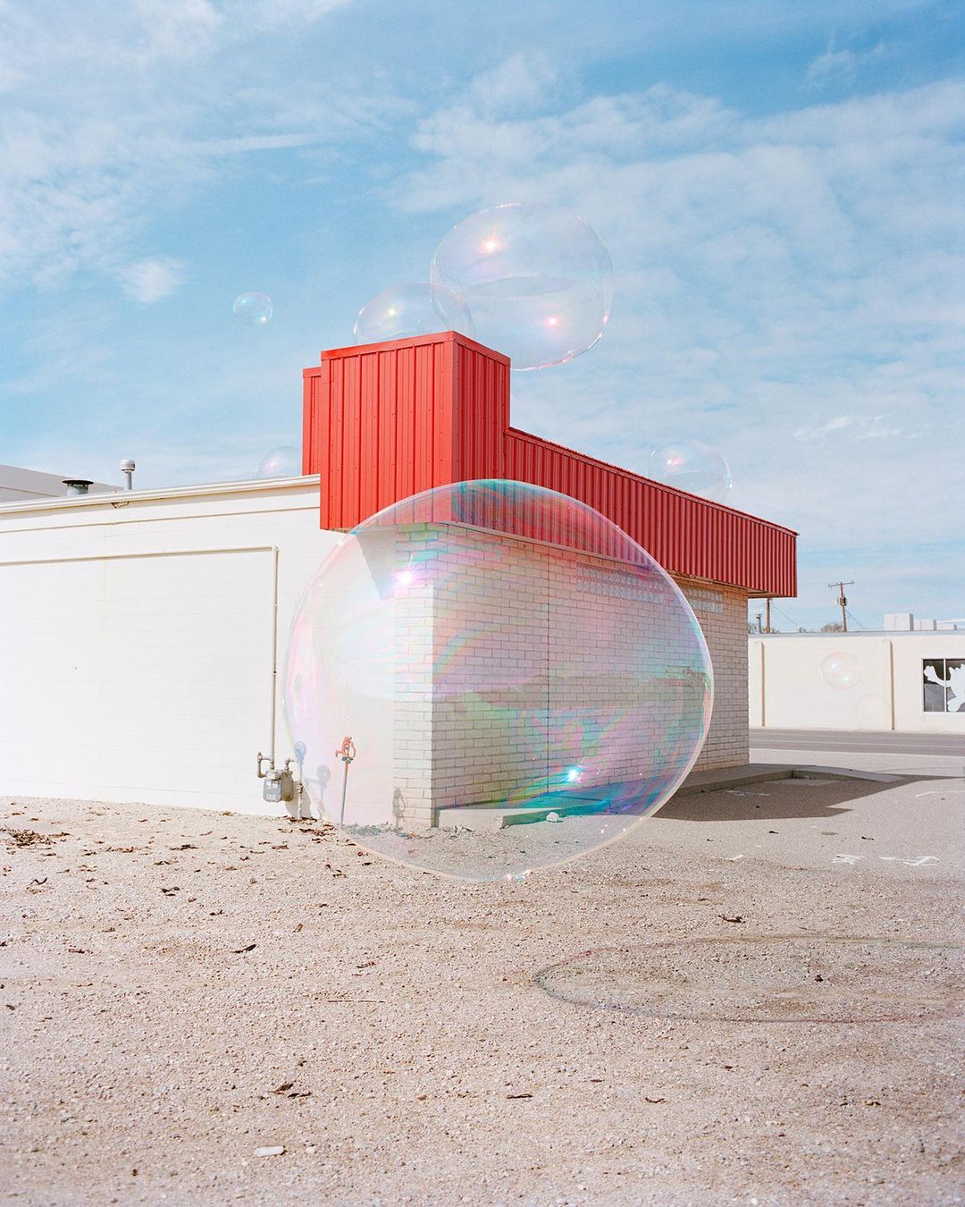 Giant transparent soap bubbles float near white and red brick buildings under a bright blue sky with scattered clouds on a sunny day.