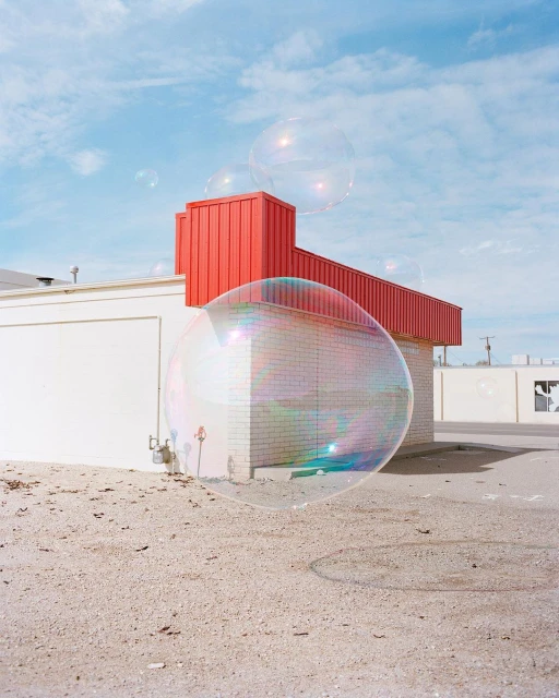 Giant transparent soap bubbles float near white and red brick buildings under a bright blue sky with scattered clouds on a sunny day.