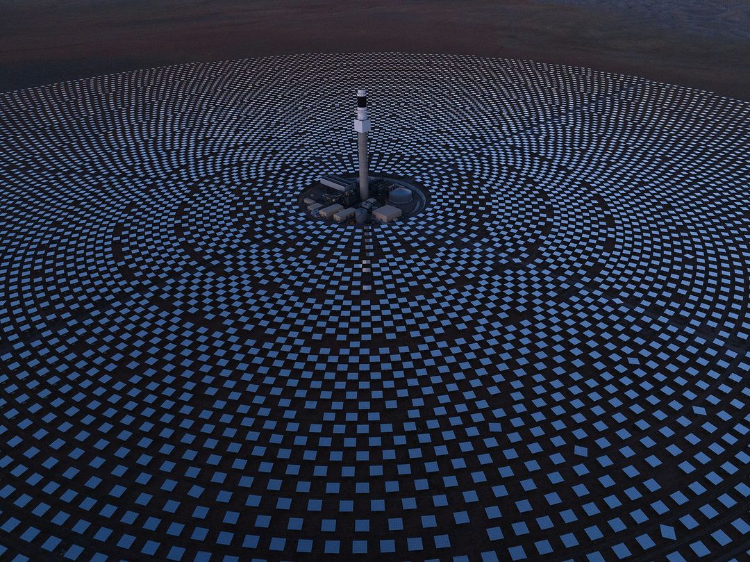 Aerial view of a solar power plant with a field of heliostats concentrating sunlight on a central receiver tower in a desert landscape.