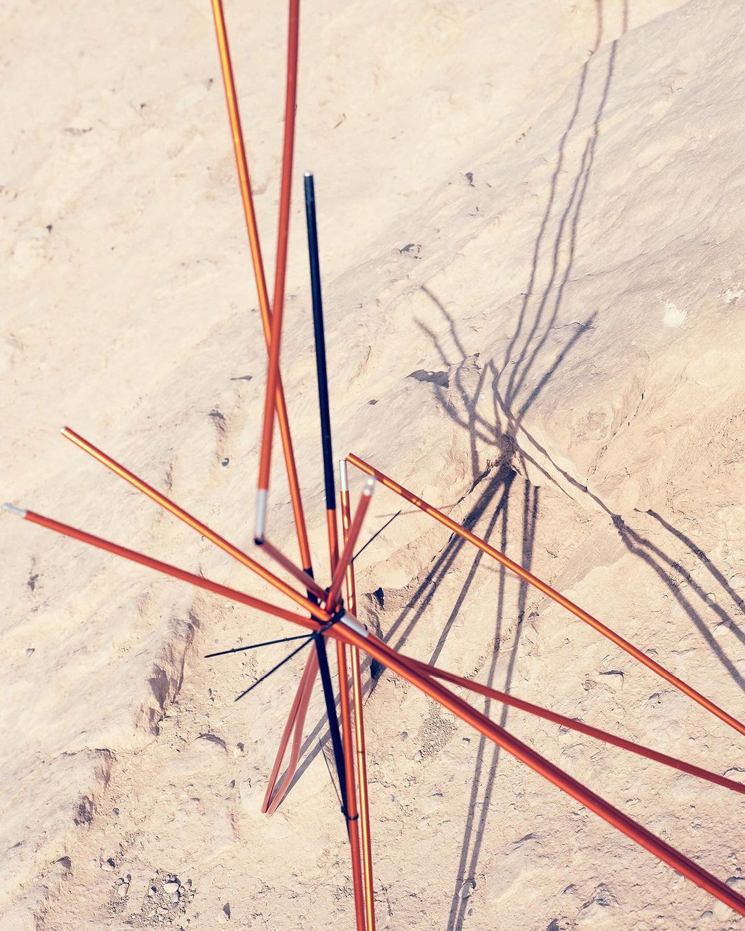 Abstract overhead shot of tent poles connected in the center, casting shadows on a tan sandy surface outdoors in full sunlight.