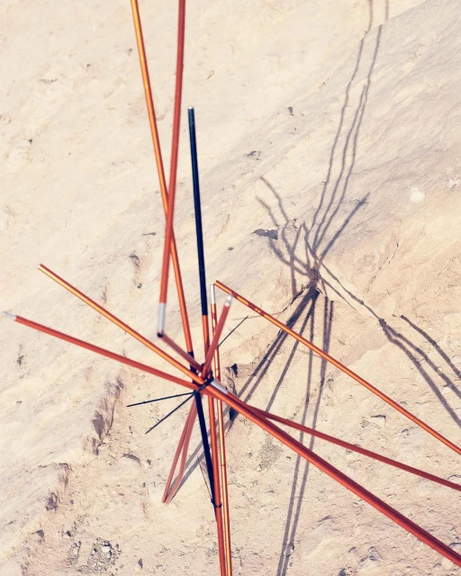 Abstract overhead shot of tent poles connected in the center, casting shadows on a tan sandy surface outdoors in full sunlight.