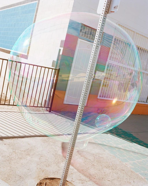 Giant bubble floats near metal sign post in an urban environment with building in the background, creating a whimsical outdoor scene.