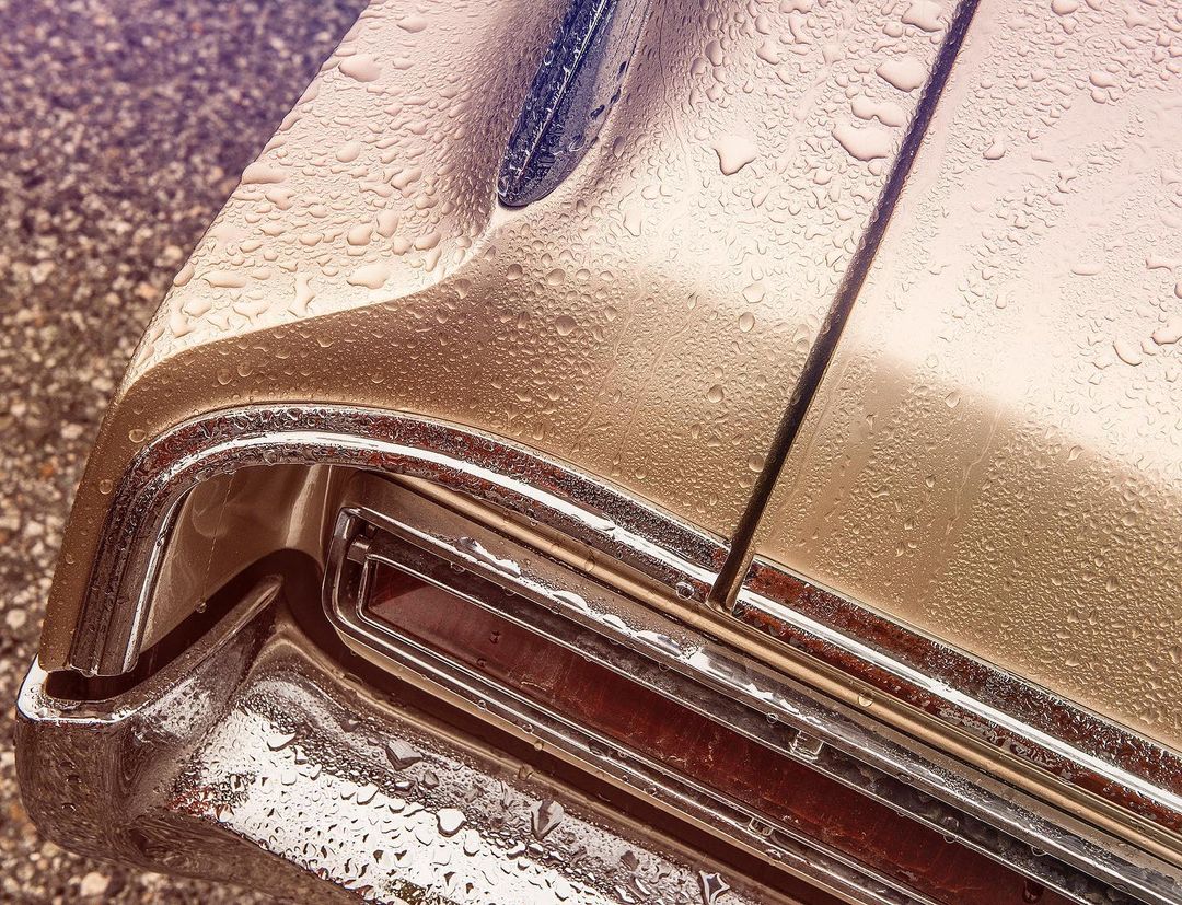 Close-up of a vintage car's rear, featuring water droplets on its golden paint, chrome trim, and taillight. A grainy asphalt surface is visible.
