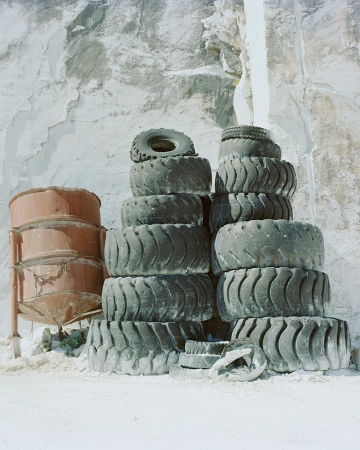 Stacked tires and rusted tanks against a textured concrete or stone wall, conveying industrial decay and minimalist composition.