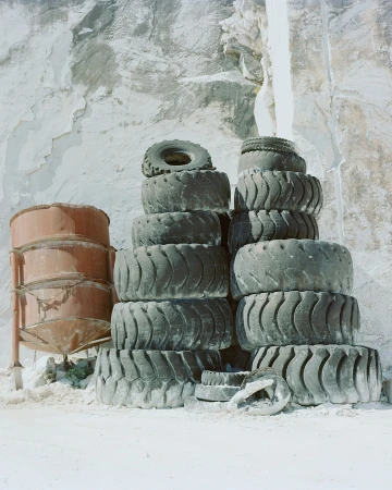 Stacked tires and rusted tanks against a textured concrete or stone wall, conveying industrial decay and minimalist composition.