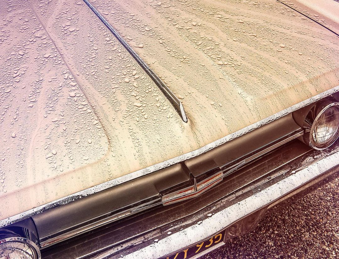 Vintage car's front hood and headlight detailed with raindrops, showcasing its metallic bumper and a glimpse of its license plate.