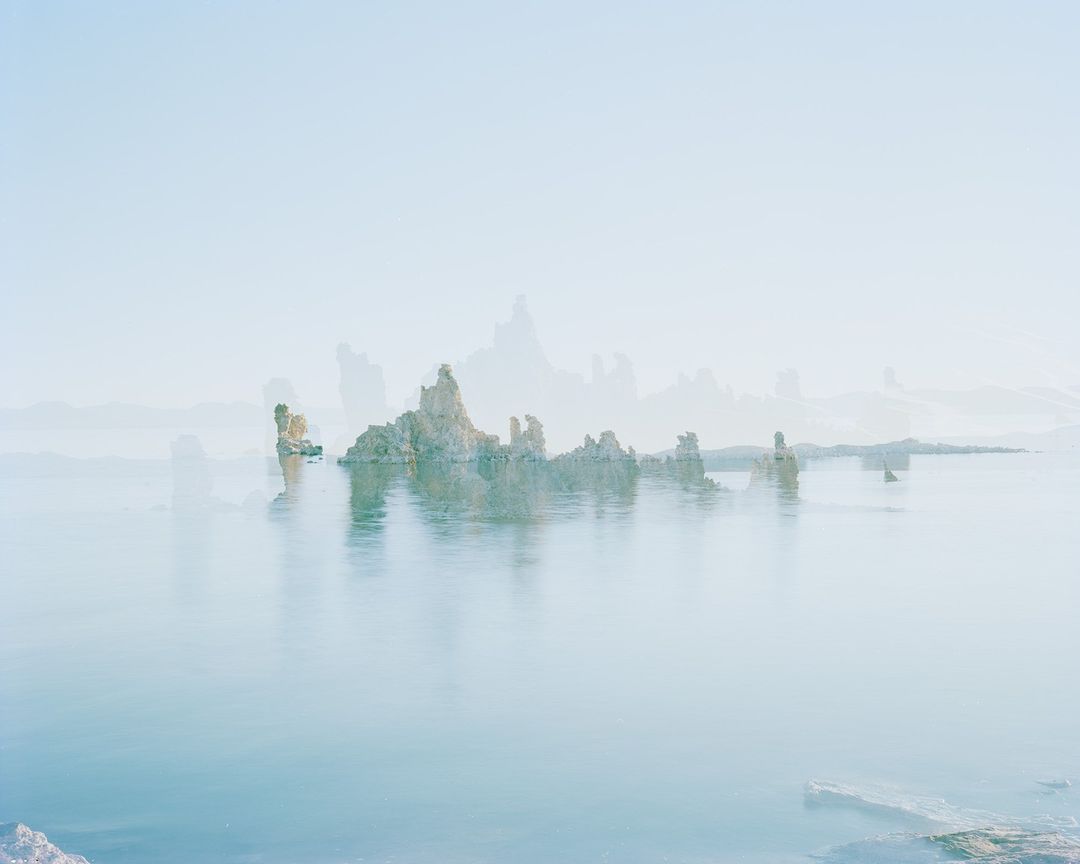 Lake landscape featuring reflective water, rock formations, and a soft, hazy atmosphere under a pale blue sky, serene and ethereal.
