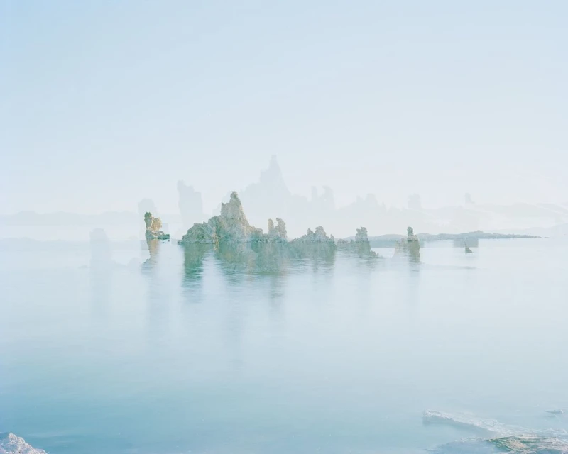Lake landscape featuring reflective water, rock formations, and a soft, hazy atmosphere under a pale blue sky, serene and ethereal.