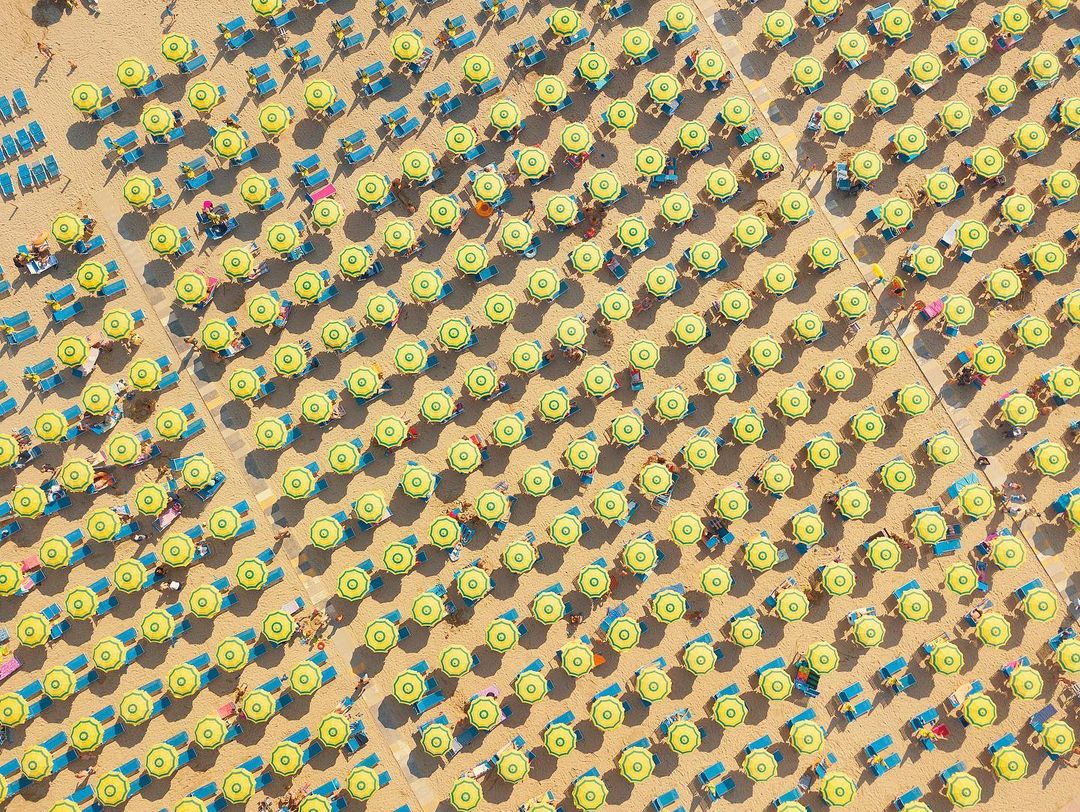 Aerial view of numerous yellow beach umbrellas and blue deck chairs evenly spaced on a sandy beach, creating a visually striking pattern.