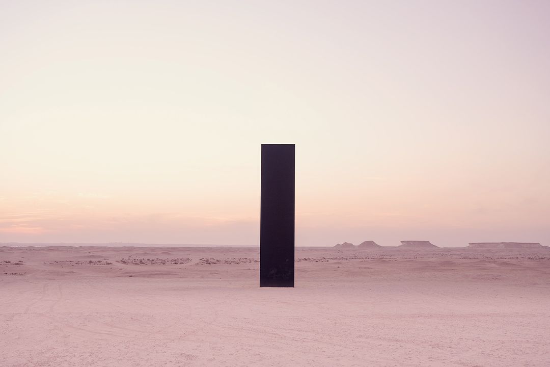 Black monolith stands in desert landscape under a pastel sky, creating a surreal, minimalist composition with geometric shapes in nature.