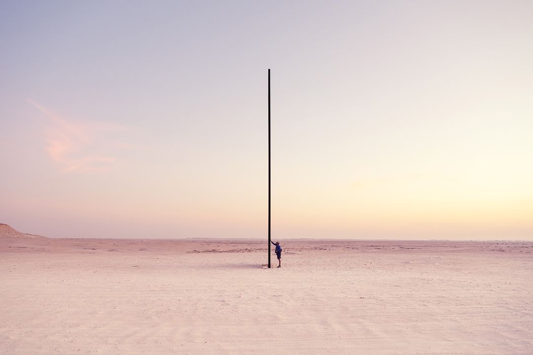 Lone figure stands beside tall, dark post in a vast, sandy desert under a soft, pastel sky at either dawn or dusk.