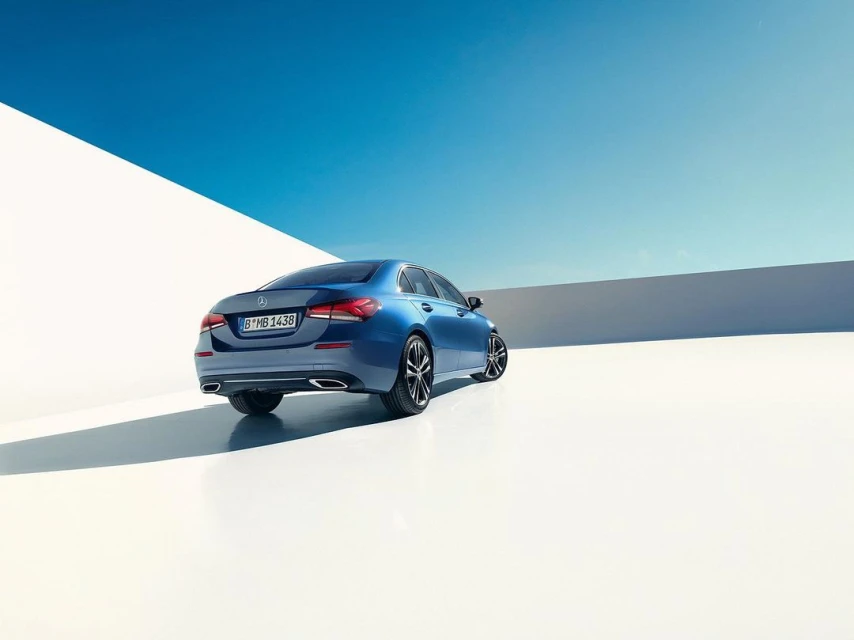 Rear view of a blue Mercedes-Benz A-Class sedan parked against a bright white wall and a clear blue sky with copy space.