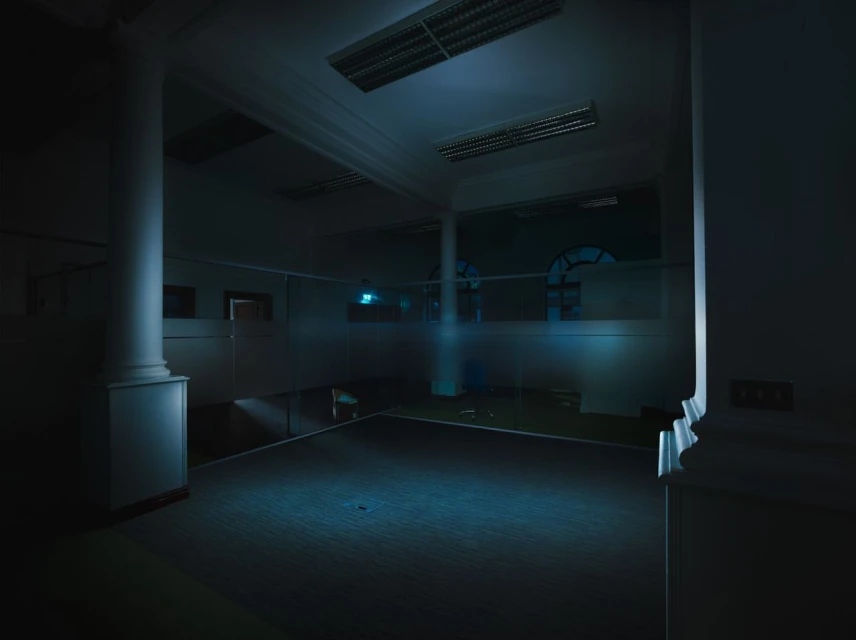 Interior shot of a large, dimly lit room with pillars, windows, and tiled floor, conveying a quiet, architectural space.