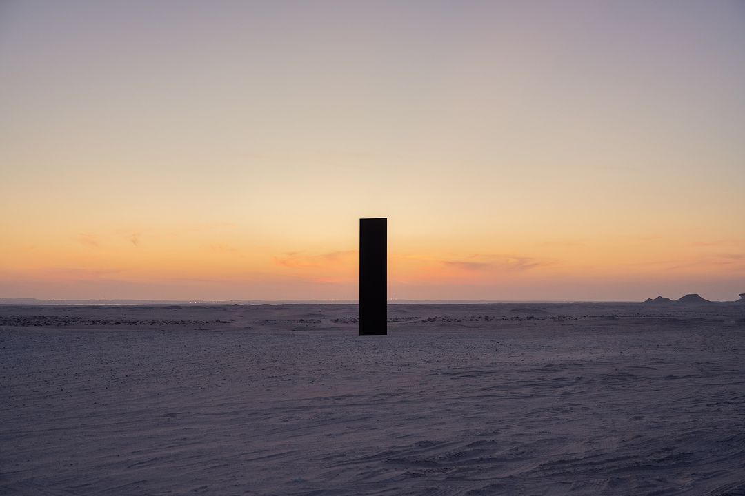 Minimalist shot of a tall, dark, rectangular monolith standing in a flat, snowy landscape under a soft, pastel-colored sunset sky.