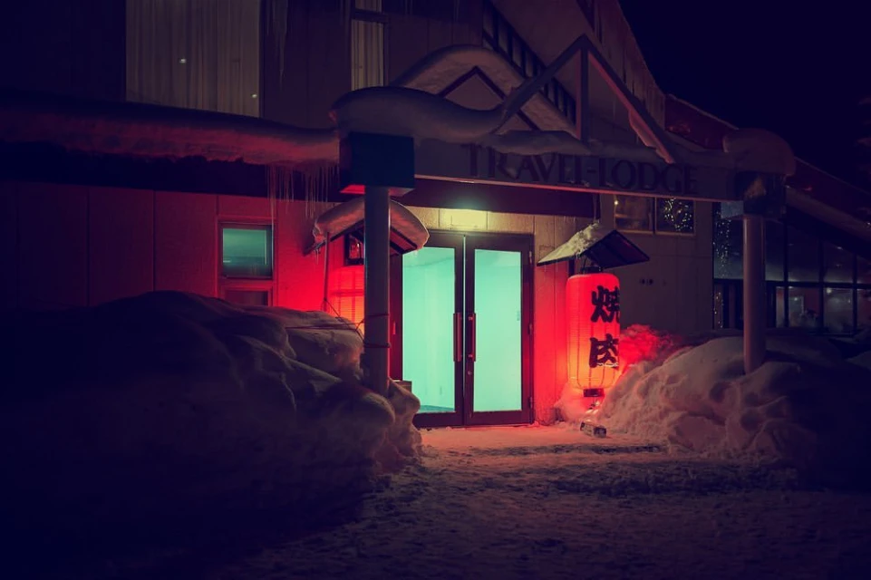 Entrance to a snow-covered lodge at night, illuminated by a red lantern. Snowdrifts surround the doorway with icicles hanging from the roof.