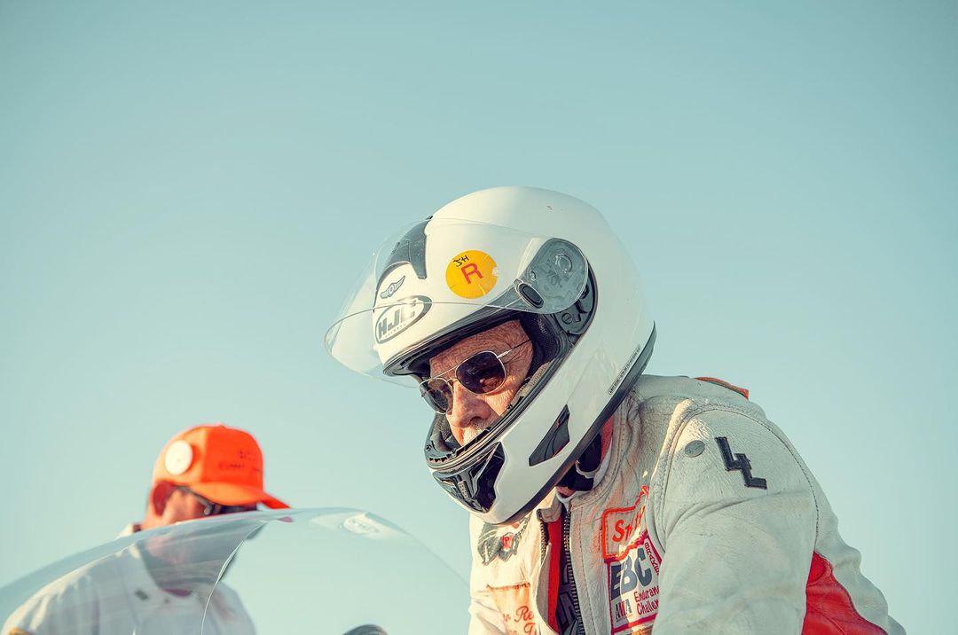 Older adult wearing racing helmet and sunglasses, standing outdoors near race vehicle on a bright, sunny day against a clear blue sky.