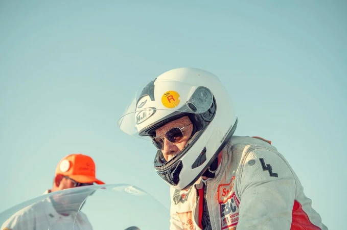 Older adult wearing racing helmet and sunglasses, standing outdoors near race vehicle on a bright, sunny day against a clear blue sky.