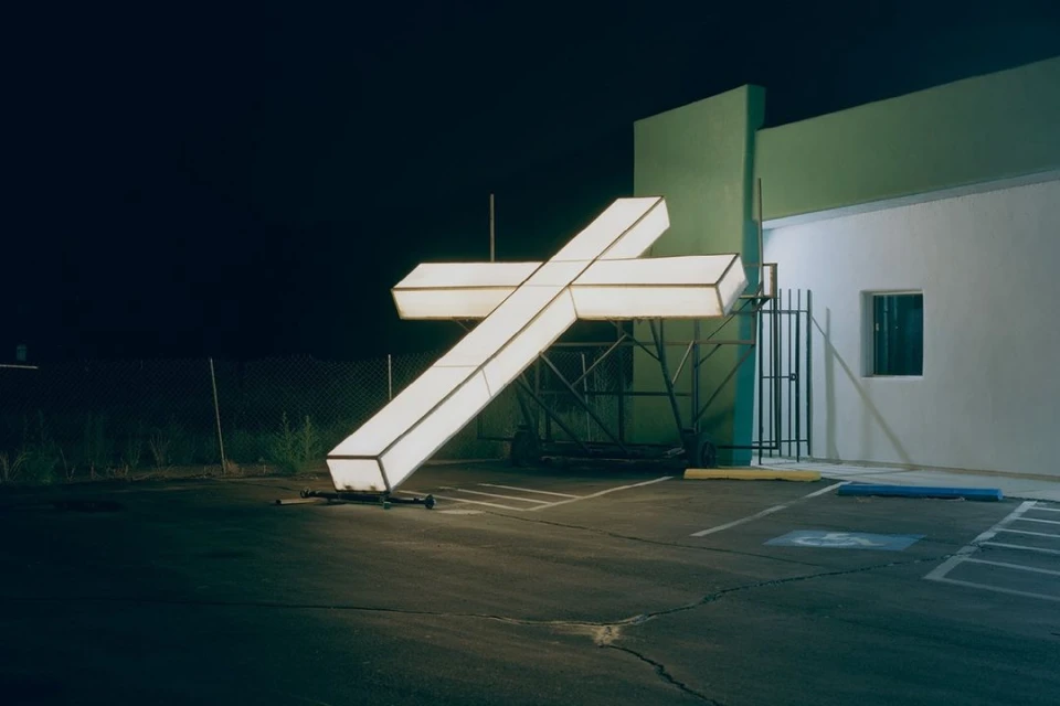 Illuminated white cross in parking lot at night, leaning against building with green accents under dark sky, no people visible.
