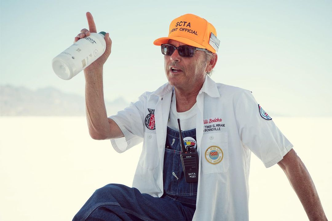 A man in work clothes and a hat points upward while holding a water bottle in an outdoor desert environment with a clear sky backdrop.