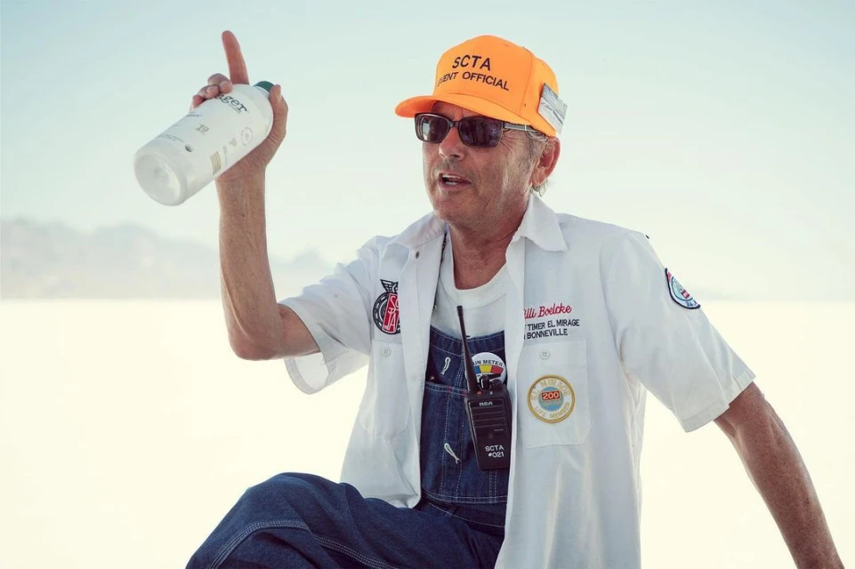A man in work clothes and a hat points upward while holding a water bottle in an outdoor desert environment with a clear sky backdrop.