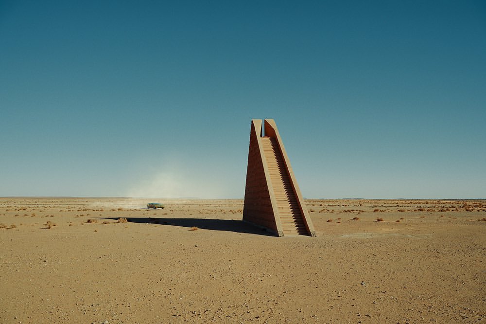 A modern art installation featuring a staircase, set in a vast desert landscape, under a clear blue sky with a distant vehicle.