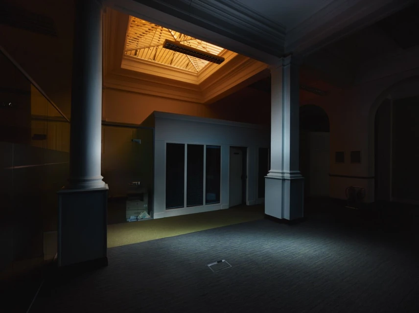 Indoor architecture showcases large columns, a windowed room, and overhead skylight, bathed in subtle illumination within a spacious interior.