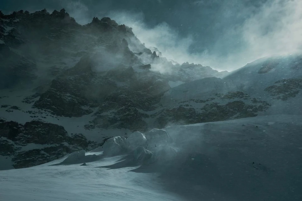 Snowy mountain landscape with rocky peaks and blowing snow on a cloudy day. Cold, dark, and dramatic winter scene.