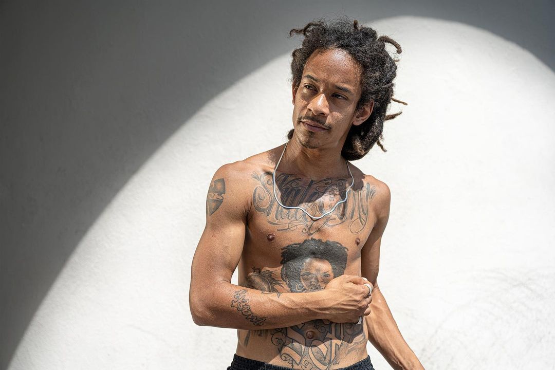 Shirtless adult male with dreadlocks and tattoos poses against a white wall with lighting effects in an eye-level portrait.