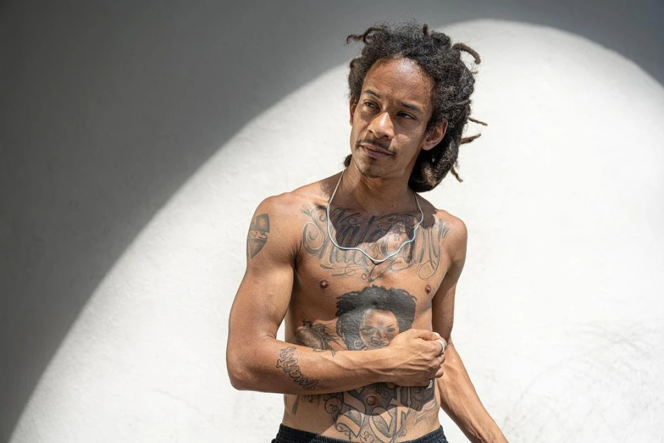 Shirtless adult male with dreadlocks and tattoos poses against a white wall with lighting effects in an eye-level portrait.