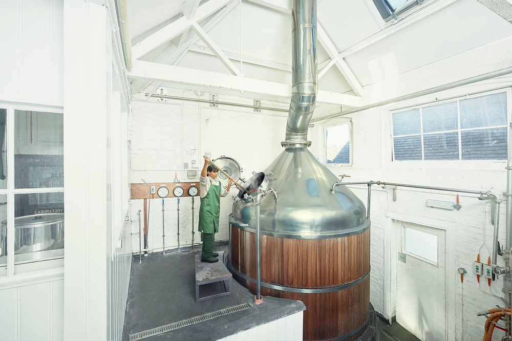 Adult male brewery worker in a green apron stands near a stainless steel brewing tank with a wooden base inside a white-walled brewery.