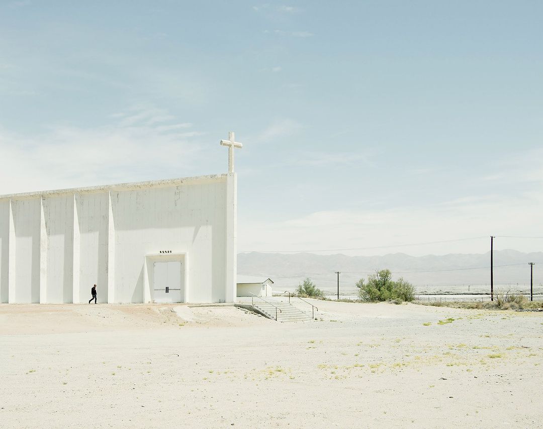 Exterior of a white concrete church with a cross, set in a desert landscape, featuring a single man walking in front of the building.