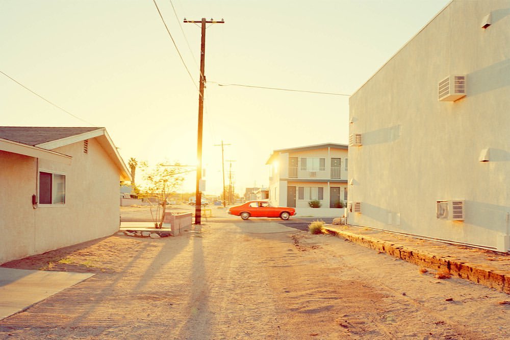 Desert street scene with vintage orange car, whitewashed buildings, and telephone pole under a bright sun, creating long shadows.