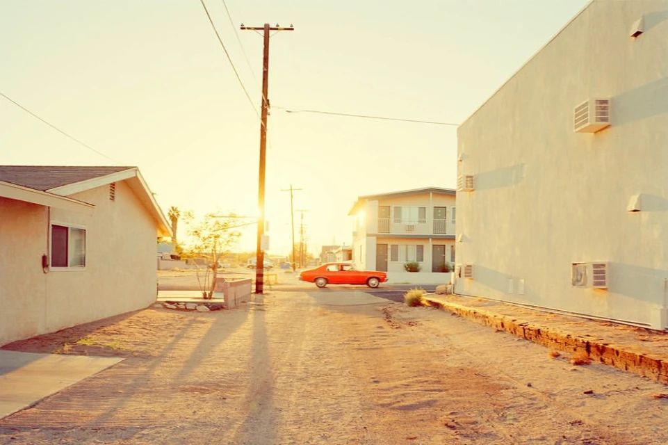 Desert street scene with vintage orange car, whitewashed buildings, and telephone pole under a bright sun, creating long shadows.