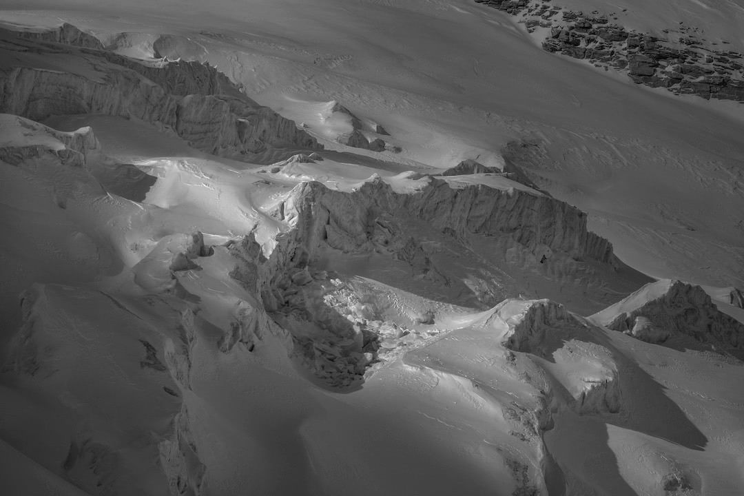 Monochromatic landscape of snow-covered mountain formations with crevasses, illuminated by sunlight, displaying a serene and stark beauty.
