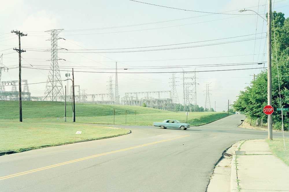 Vintage car on a road near power lines and towers, set against a pastel sky with green grass and trees in an American suburb.