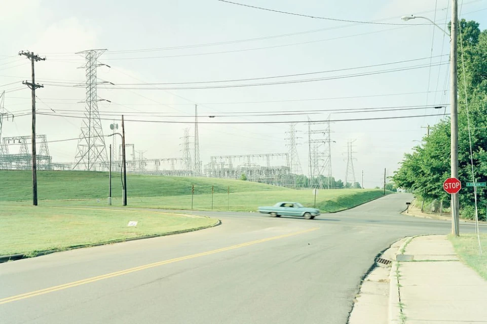 Vintage car on a road near power lines and towers, set against a pastel sky with green grass and trees in an American suburb.