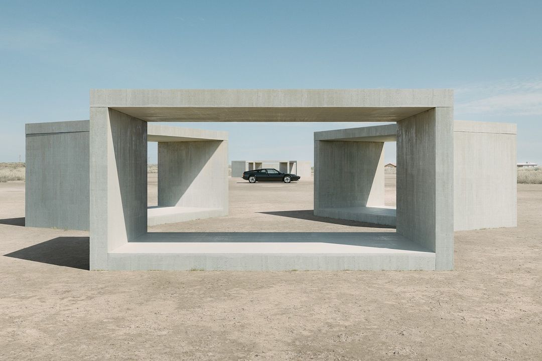 Vintage car parked in the distance, framed by concrete geometric structures in a desert landscape under a clear blue sky. Automotive photography.