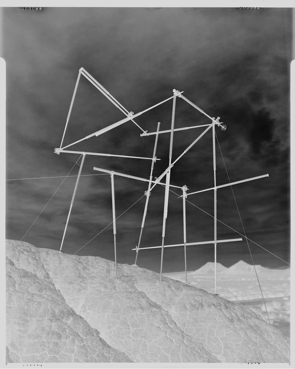 Abstract construction on a rocky hill under a cloudy sky. Complex geometric scaffolding structure contrasts with the natural landscape.