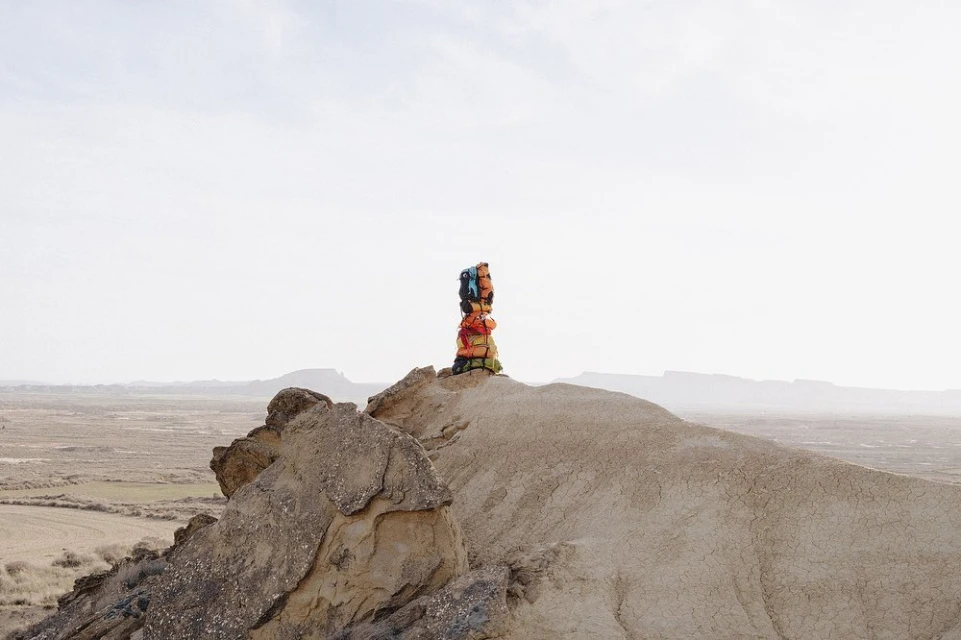 Person stacks colorful luggage bags on a rock formation in the desert, captured with a wide shot under a bright sky, creating a travel image.