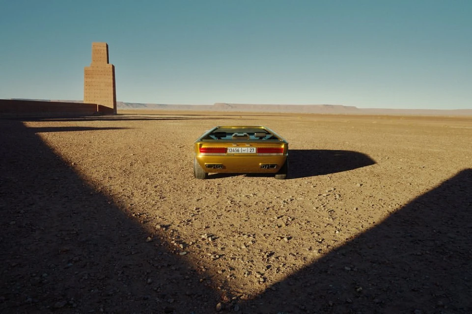 Gold sportscar parked in a desert landscape with a blue sky, featuring a geometric brick structure in the background.